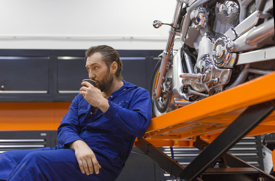 A man in the workshop sits near a motorcycle and drinking coffee.
