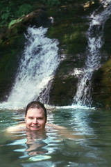 young smiling man swimming in lake with waterfall on background