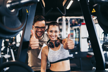 Young fit and attractive woman working out in modern gym together with her personal fitness instructor or coach. They looking at camera and smiling while showing thumb up.