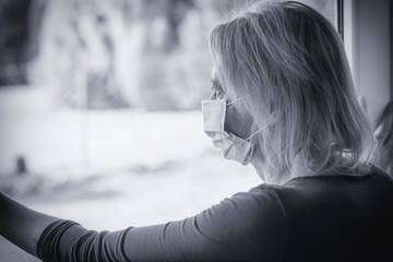 Lonely woman in a quarantine with a disposable protective face mask looks sadly out of the window. 