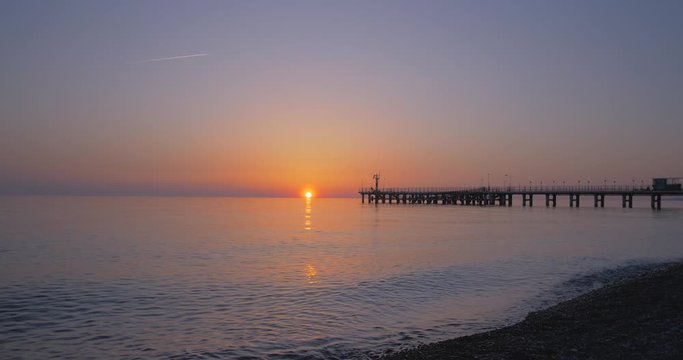 Sunset on the seashore with a pier and a plane flying by in calm windless weather.