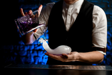 Close-up bartender gently pouring drink from glass mixing cup into smoky glass flask of unusual...