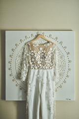 Beautiful off white wedding dress hanging on a wooden hanger against a cream-colored wall.