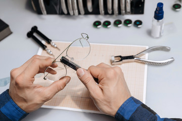 Young professional optical technician is fixing the bridge of eyeglasses in workshop with special pliers. Eyesight concept.