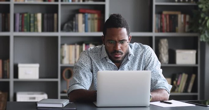 Thoughtful young african american man in eyeglasses watching educational lecture online on computer, writing notes on paper, web surfing searching additional information in internet, sitting at home.