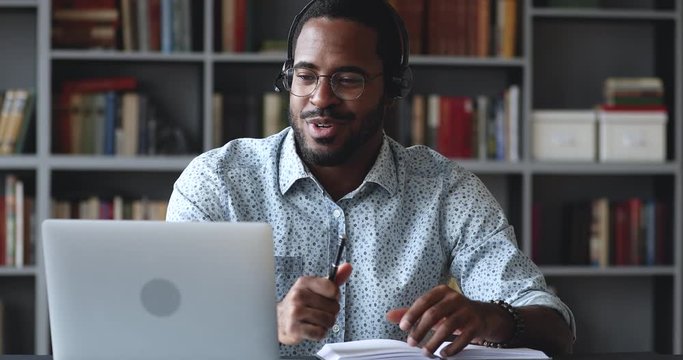 Happy young african american guy in glasses wearing wireless headphones with microphone, looking at laptop, holding video call talking speaking with consultant teacher mentor coach, writing notes.