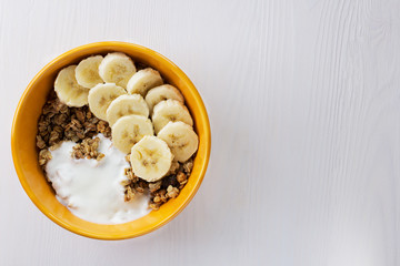 The muesli with banana and yogurt in a bowl on the table on a white wooden background.