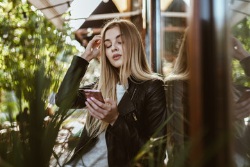 a proud beauty with long blonde hair is standing at the cafe and lowering her eyes straightens her hair with her hand in her other hand the phone