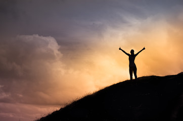 Happy female standing on a hill facing the sunset putting her arms, thumbs up feeling happy,...