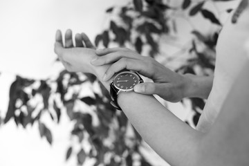 close up photo of female hand wearing black wrist watch  with arabic numerals with green flower in background. black and white photo. 