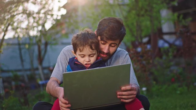 Good time young dad with big chin and his small son enjoying the time in the garden they playing a game on the laptop and feeling very excited