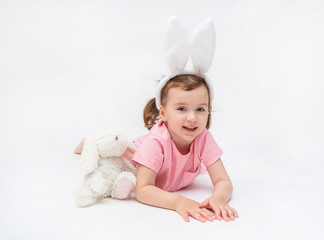 A cute girl is lying on her stomach on a white background with an empty seat. A girl with hare ears and a stuffed rabbit toy. Little girl in a pink t-shirt. The girl looks at the camera and smiles