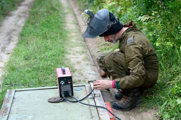 Old man welder in brown uniform, welding mask and welders leathers, weld metal door with arc...