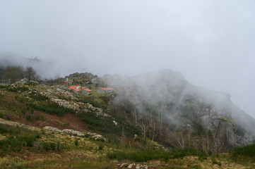 Small houses with orange roofs in the middle of hills. Green grass contrasting with yellow grass. Foggy day. Freita mountain range.