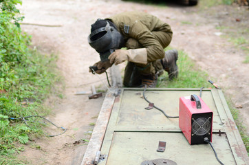 Old man welder in brown uniform, welding mask and welders leathers, weld metal door with arc...