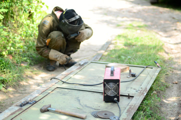 Old man welder in brown uniform, welding mask and welders leathers, weld metal door with arc...