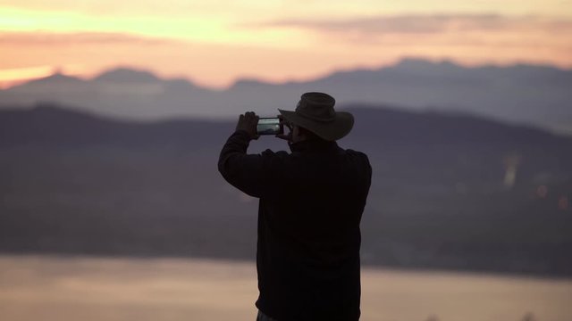 Lockdown shot from the back of a man wearing hat taking pictures with his phone of a scenic bay Amon mountains at dusk - Cape Town, South Africa