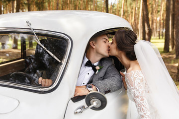 Elegant newlyweds, groom and bride kisses near white Just Married car at wedding day