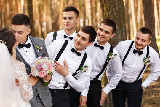 Bride and groom kisses near friends, groomsmen with bow ties and suspender looks at newlyweds at wedding day