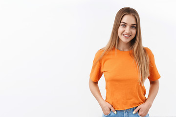 Obraz premium Portrait of gorgeous female student, blonde girl in orange t-shirt, smiling camera enthusiastic, with lovely happy expression standing over white background, relaxed pose with hands in pockets