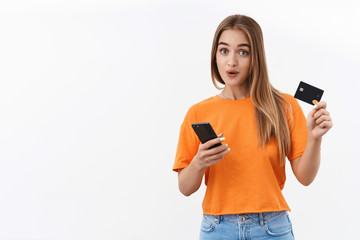 Portrait of intrigued and surprised blond girl found awesome delivery service, good online shop trying make internet purchase, order with credit card, holding mobile phone and look curious camera