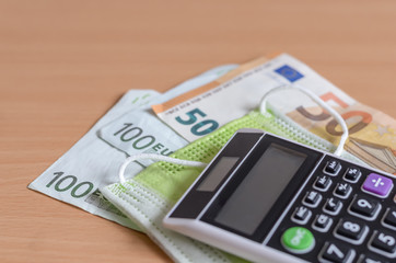 Close-up  of  euros banknotes   and  medical disposable  face mask   with   handheld mini calculator  on wooden background . The economic impact of coronavirus  in Europe .