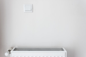 White radiator on white wall with light switch in home indoor. Modern home comfort concept.