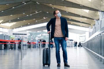 Full length shot of man passeger with suitcase in international airport wears protective face mask to avoid coronavirus infection. Male with luggage going to travel abroad. Epidemic, health care