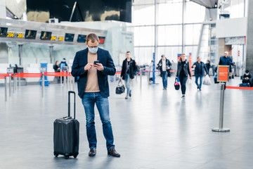 Horizontal shot of man poses in crowded airport, uses cell phone, checks time of flight online, stands near suitcase, wears medical mask during coronavirus crisis. Health care, traveling concept