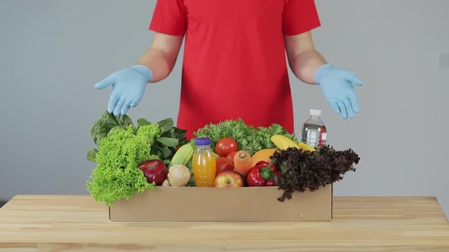 Online shopping concept. serious Male courier in red uniform, protective mask and gloves with a grocery box with fresh fruits and vegetables. Home delivery food during quarantine coronavirus