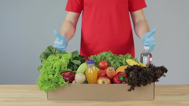 Male courier in red uniform, protective mask and gloves with a grocery box with fresh fruits and vegetables showing thumbs up. Home delivery food during quarantine coronavirus
