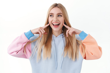 Naklejka premium Beauty, lifestyle and fashion concept. Upbeat good-looking blond girl poking her cheeks and smiling, looking away with joyful, happy emotion, promote skincare cosmetics or makeup