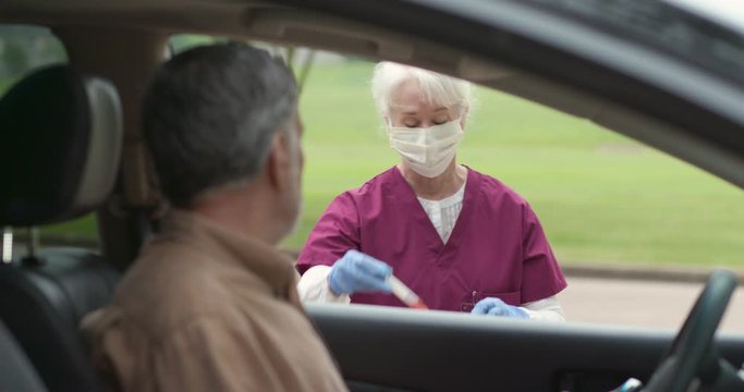 A mature man at a mobile testing location who has finished a do it yourself swabbing procedure watches a healthcare worker process the vial that he has placed on a table outside the vehicle.