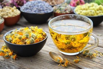 Cup of healthy marigold tea and calendula flowers in bowl. Medicinal herbs - lavender, marigold, rose, daisies, Helichrysum on background. Herbal medicine. - obrazy, fototapety, plakaty