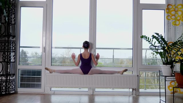 Young ballet dancer, teenage girl performing exercise twine , stretching her legs, sit in twine, against the background of a large panoramic window.