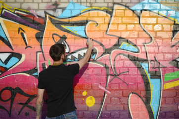 Young graffiti artist with backpack and gas mask on his neck paints colorful graffiti in pink tones on brick wall. Street art and contemporary painting process