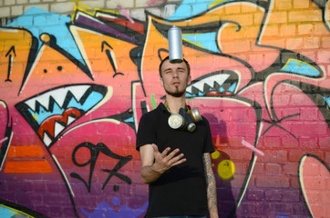 Young graffiti artist with gas mask on his neck throw his spray can against colorful pink graffiti on brick wall. Street art and contemporary painting process
