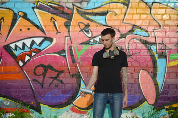 Young caucasian graffiti artist in black t-shirt with silver aerosol spray can near colorful graffiti in pink tones on brick wall. Street art and contemporary painting process