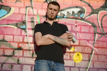 Young caucasian graffiti artist in black t-shirt with silver aerosol spray can near colorful graffiti in pink tones on brick wall. Street art and contemporary painting process