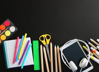 Top view of school stationery: colored pencils, paints, crayons, markers, ruler, notepad, electronic tablet and headphones on a background of black wooden table. Back to school flat lay. Copy space