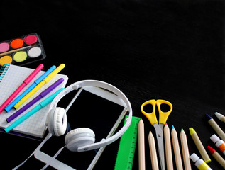 Top view of school stationery: colored pencils, paints, crayons, markers, ruler, notepad, electronic tablet and headphones on a background of black wooden table. Back to school flat lay. Copy space