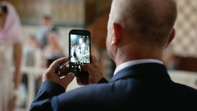 Close-up. Old man with a bald head in a suit takes pictures on the phone in a church.