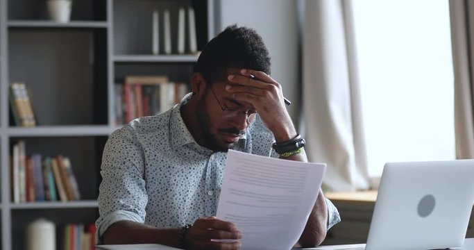 Thoughtful young african man looking through reading paper document, feeling stressed about banking loan money debt notification. Depressed biracial guy frustrated about high bills, dismissal notice.