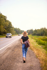 Fototapeta premium Blonde woman wearing a hat walking along the side of a road with a backpack.