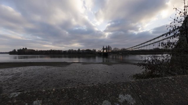 The camera tracks upwards and pans right as the sun sets over the Hammersmith Bridge in the London borough of Hammersmith. Lovely clouds stream overhead as the camera reveals more of the bridge