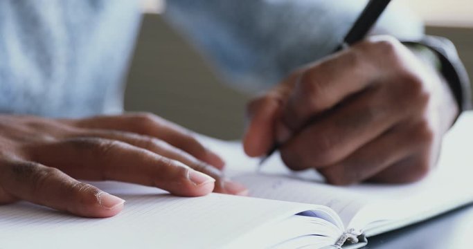 Close up young african american male student holding pen, writing notes in paper notepad. Confident mixed race businessman planning workday, noting daily cases in organizer before workday start.