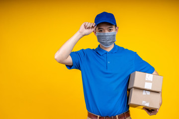 Obraz na płótnie Canvas Young Asian handsome delivery man wearing mask holding boxs on left arm and touching his cap over yellow isolate background. Work from home and delivery concept.