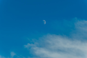 A wide angle minimalist shot of the crescent moon against the background of the blue sky with some feather clouds in the late afternoon