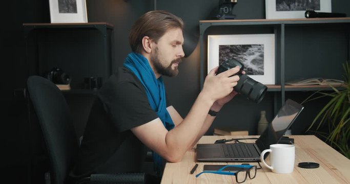 Mature bearded man in black t-shirt and blue scarf reviewing and picking photos for retouching. Photographer using digital camera and laptop for working in studio.