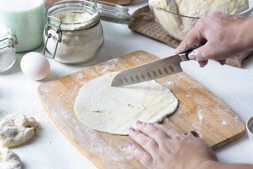 A woman cuts a flat circle of dough on a wooden board, next to flour, eggs, milk. Russian kitchen